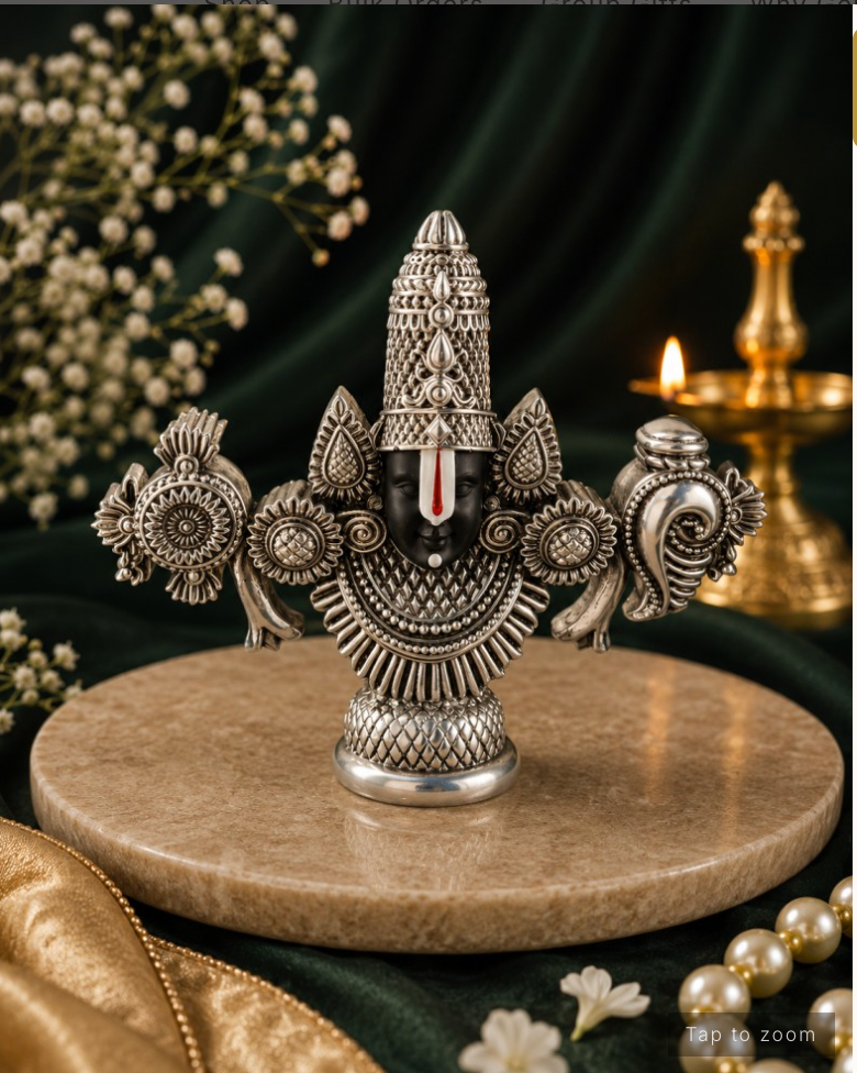 An ornate silver-toned deity bust sculpture displayed on a round beige stone plinth, with dark green draped fabric, small white flowers, and a lit brass oil lamp in the background.